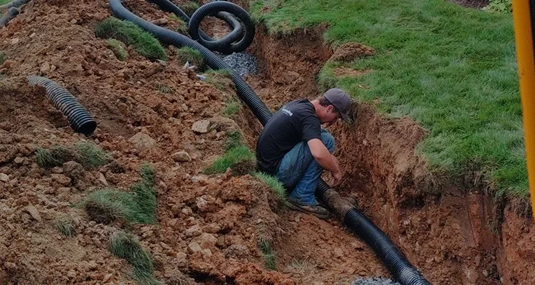 A worker installing drainage pipes in a freshly dug trench in a yard.